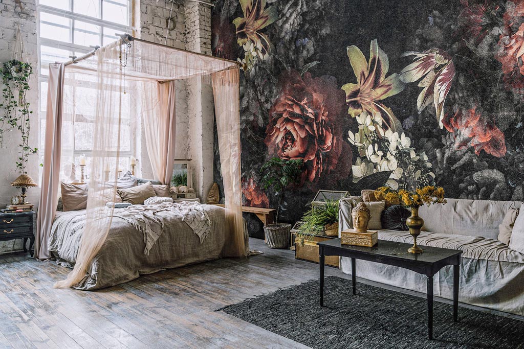 Bed with baldachin and huge window behind it, couch and black table with bust of Buddha and flower vase in Bohemian style room. Gray carpet on wooden floor and brick wall with empty picture frames
